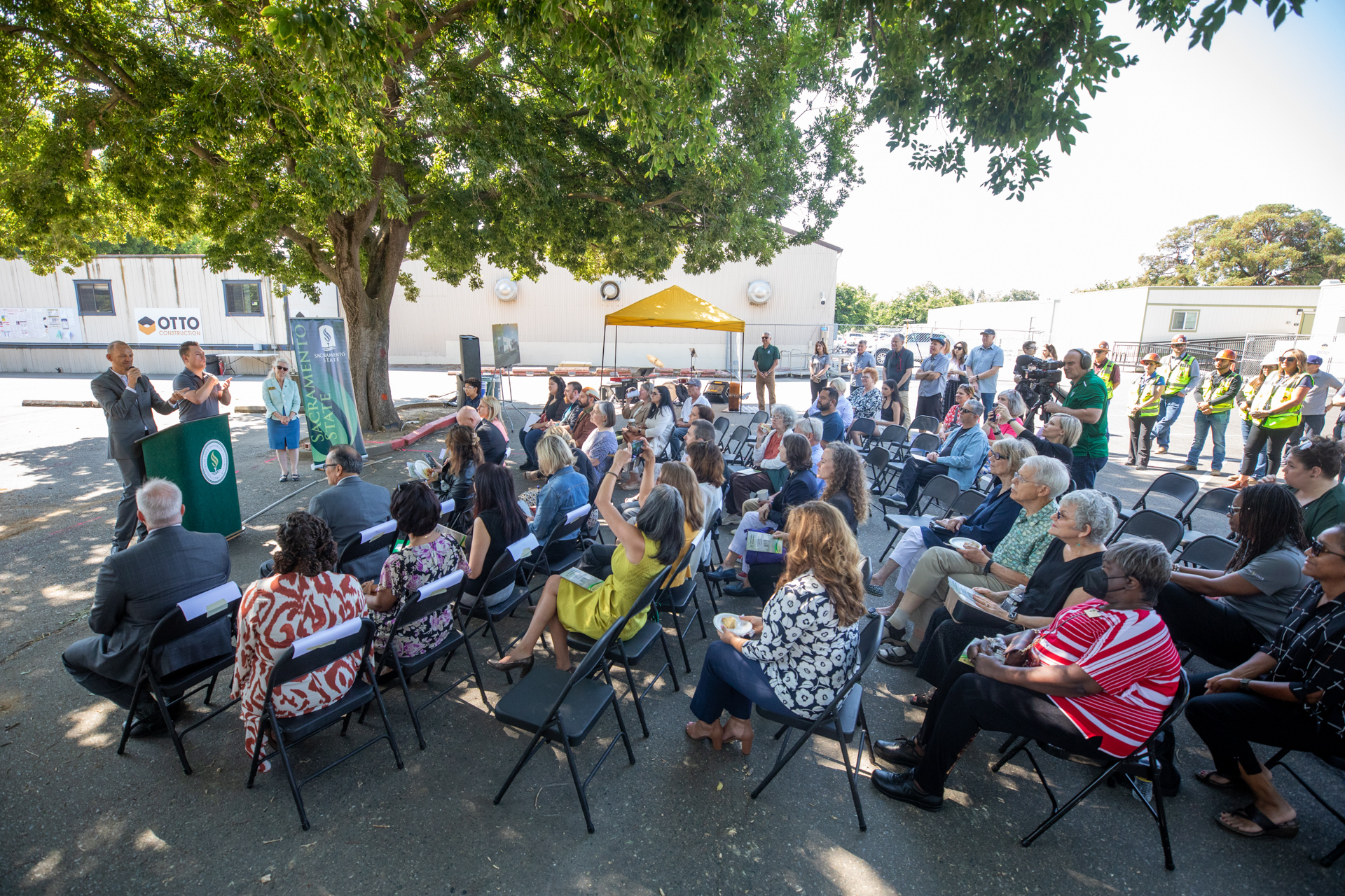 Attendees sit in share under a shade tree while a speaker talks at a groundbreaking event.