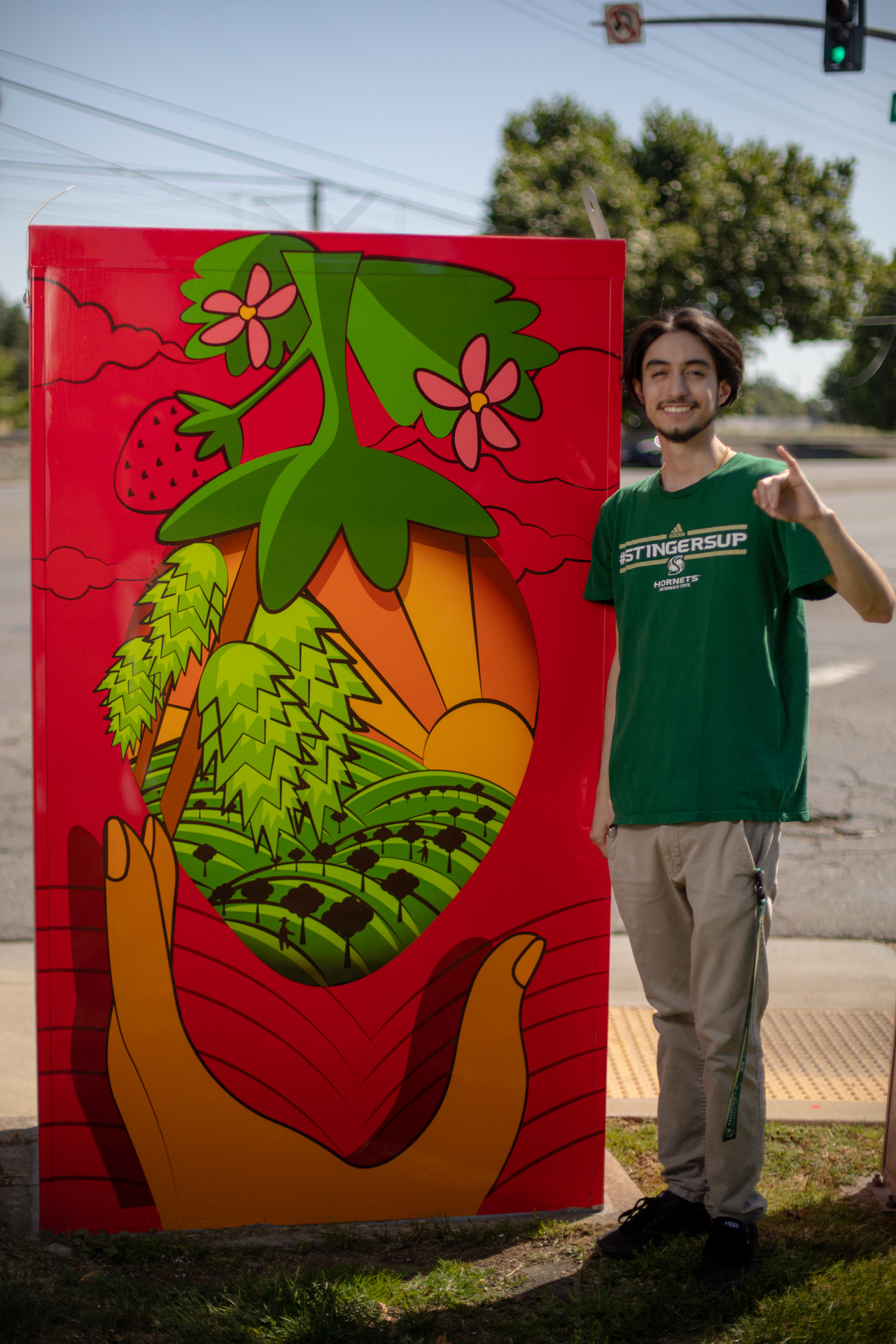 A Graphic Design graduate salutes Stingers Up with one pinky in the air next to an art-wrapped utility box he helped design.