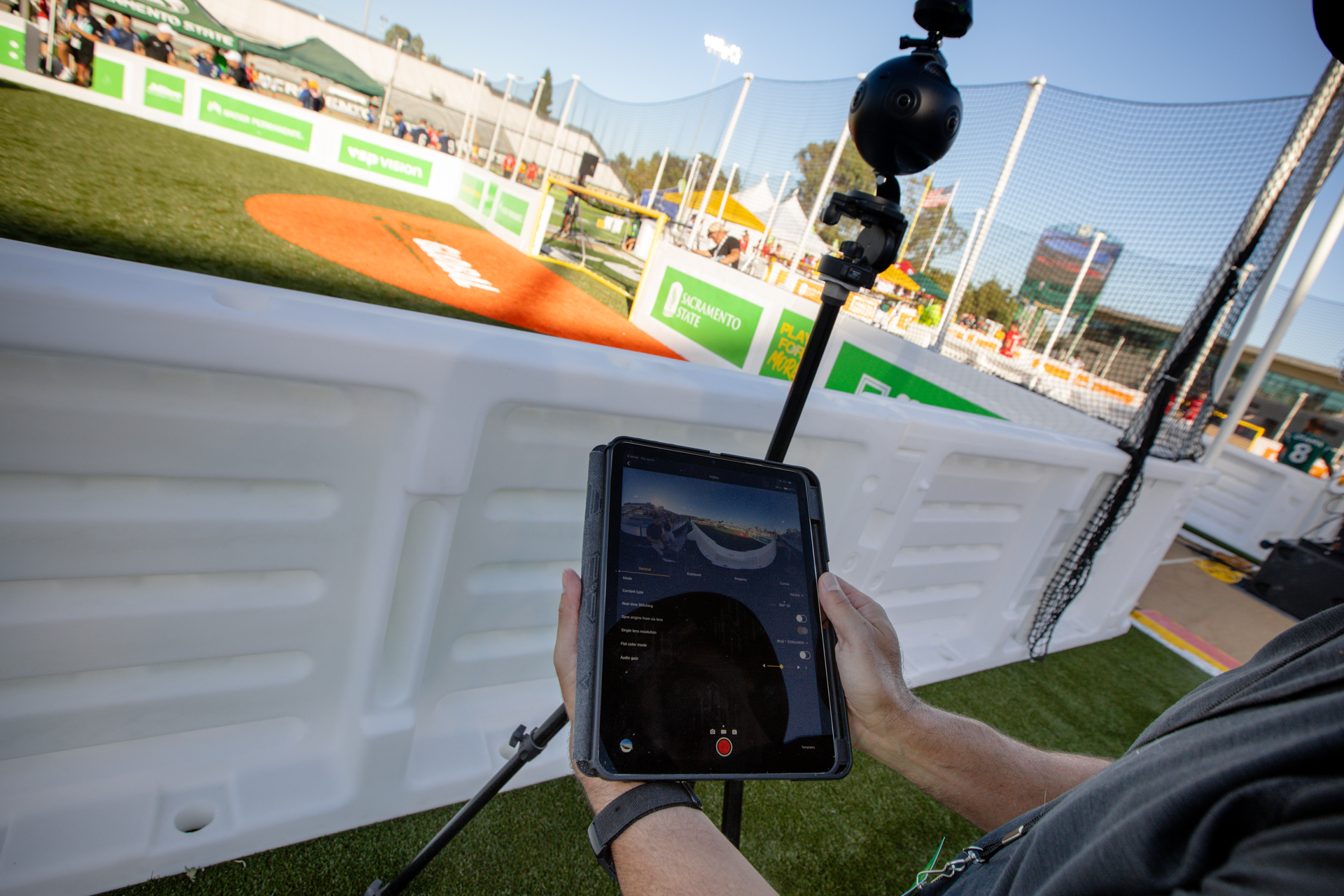 Two hands holding a tablet computer, next to a 3D camera on a tripod, with street soccer fields in the background