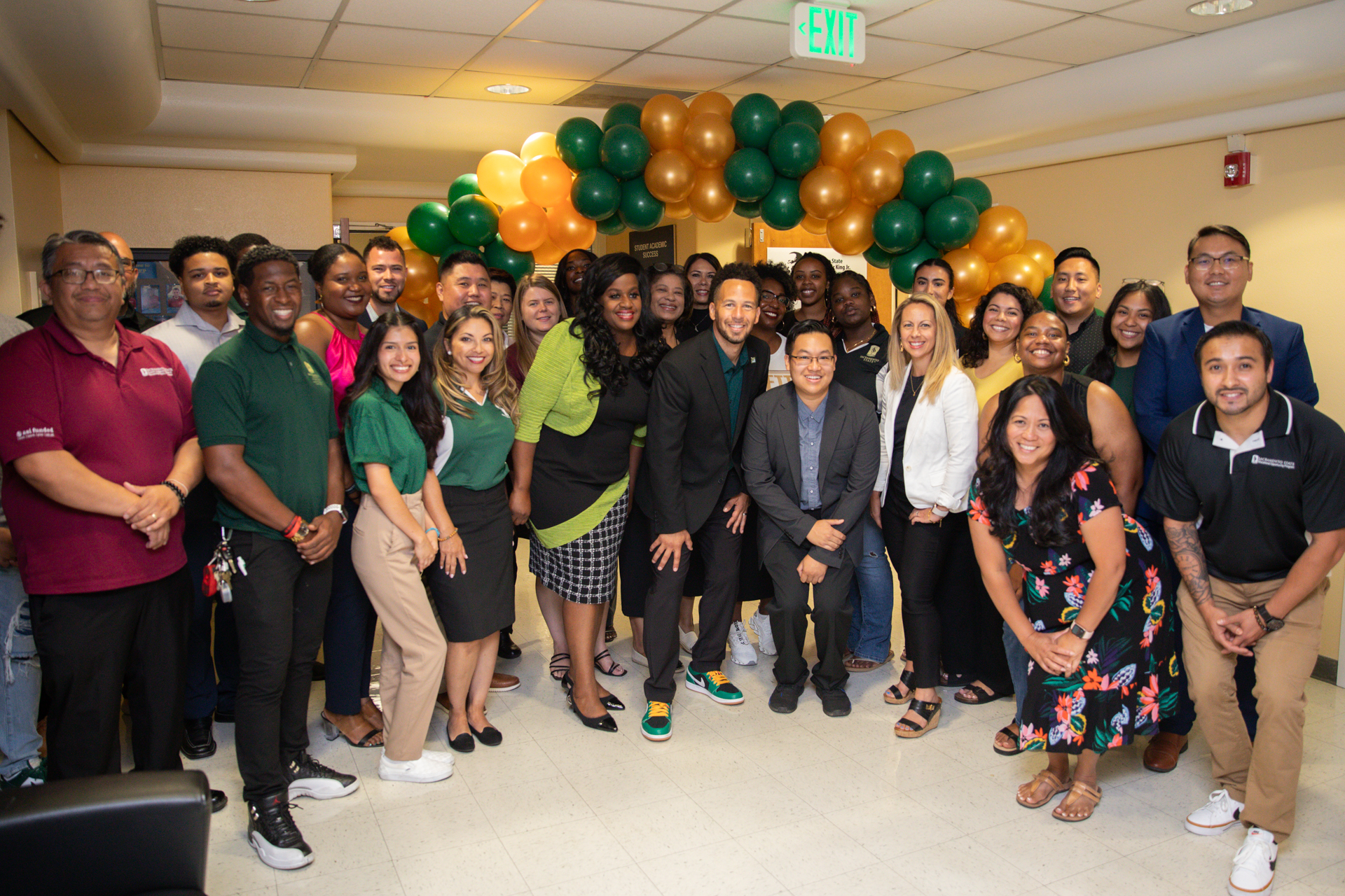 President Luke Wood poses for a photo with students and staff, beneath a green and gold balloon arch.