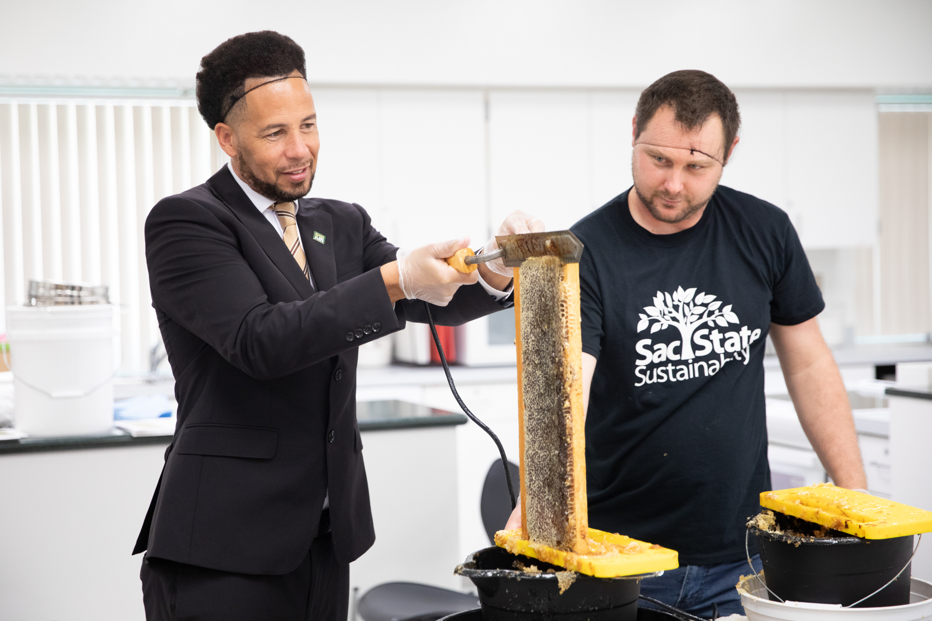 President Luke Wood and a man in a Sac State Sustainability T-shirt scrape honey off of a comb.