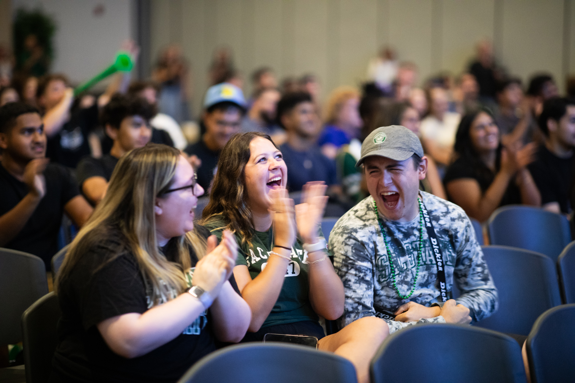 Three individuals sitting in the University Union Ballroom, cheering during a football watch party.