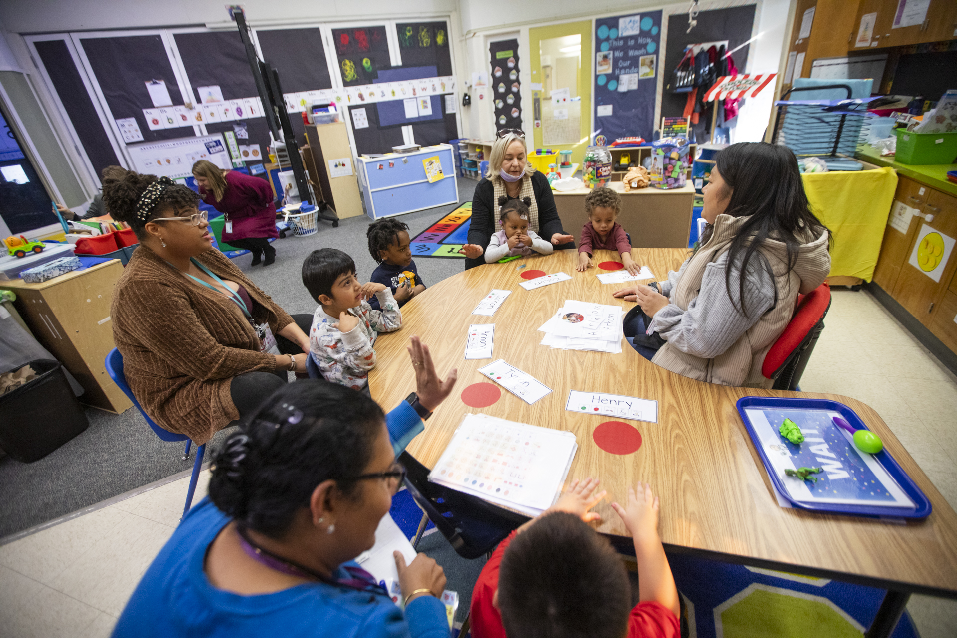 Three women sit at a table with preschool students inside a classroom.