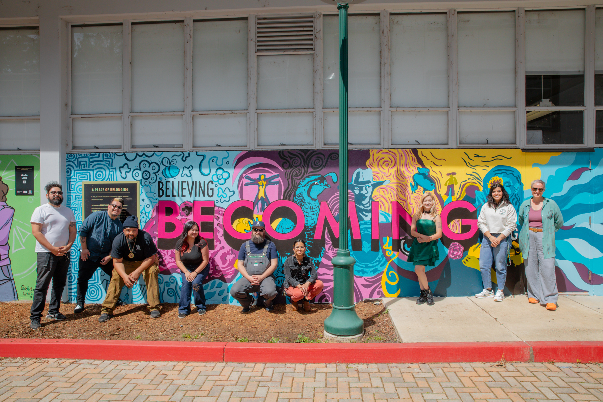 Nine artists pose for a photo, standing outdoors in front of the "Becoming" mural.