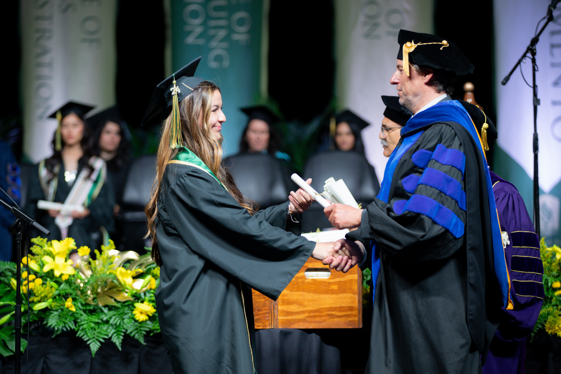 A woman dressed in academic regalia shakes the hand of a University dean while receiving a diploma during Commencement.