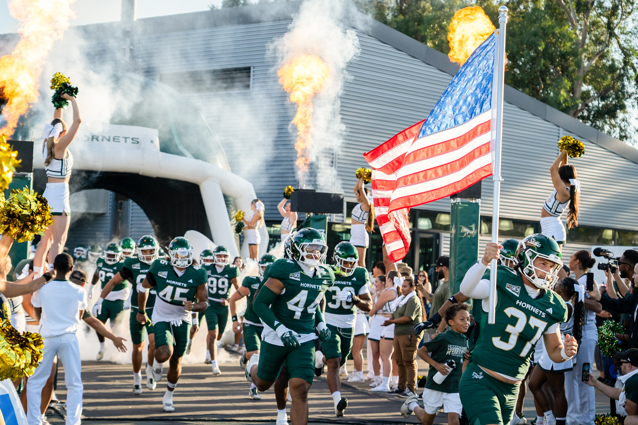 Football players run through giant, inflatable helmet with one carrying the American flag as cheerleaders wave amidst smoke.