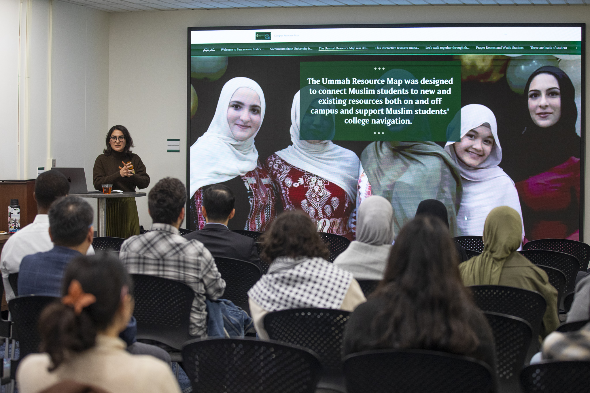 A woman at a podium demonstrate new interactive maps on a large screen covering for an audience.