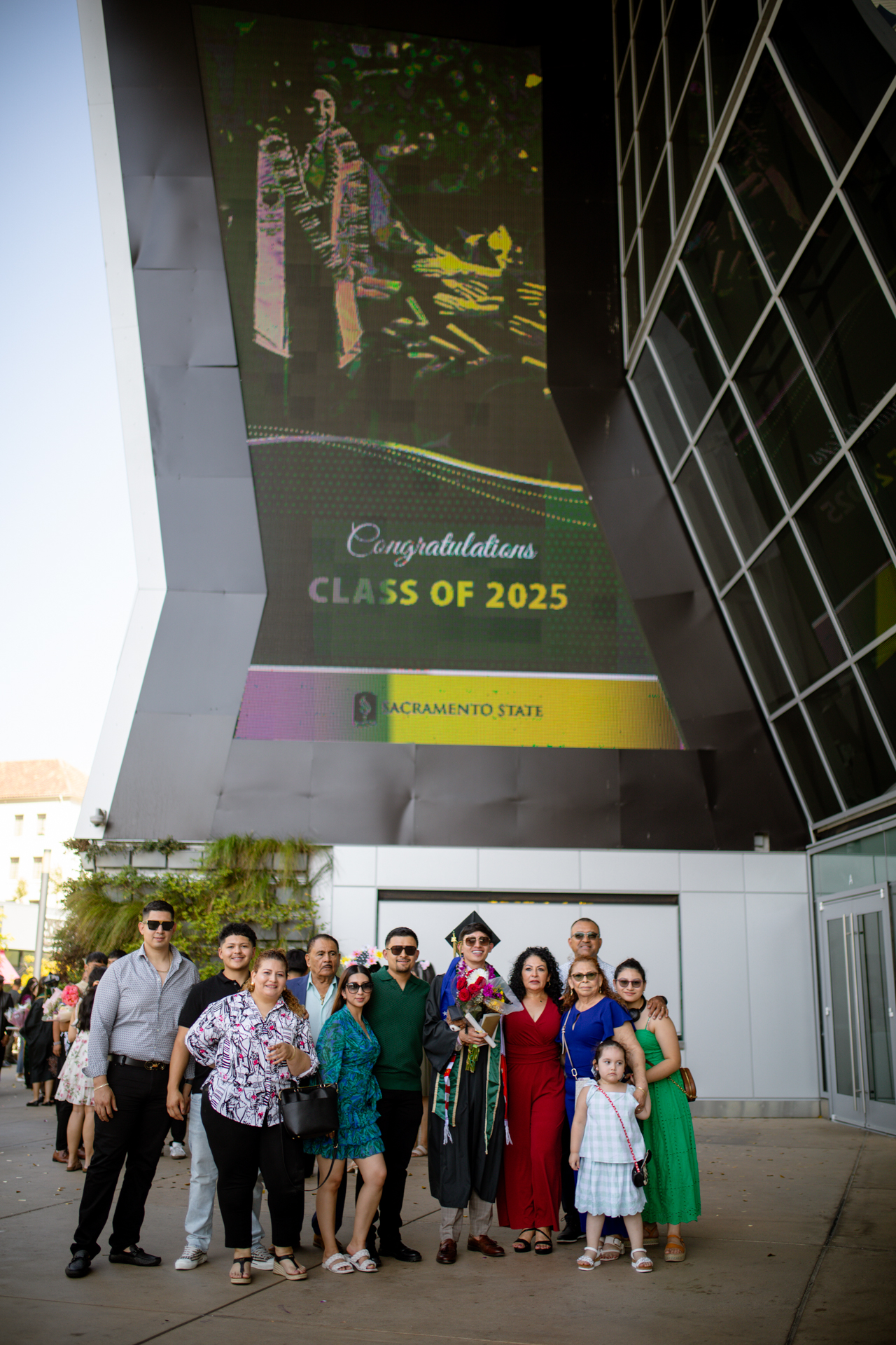 A family and their graduate pose for a photo outside Golden 1 Center near a Commencement digital sign.