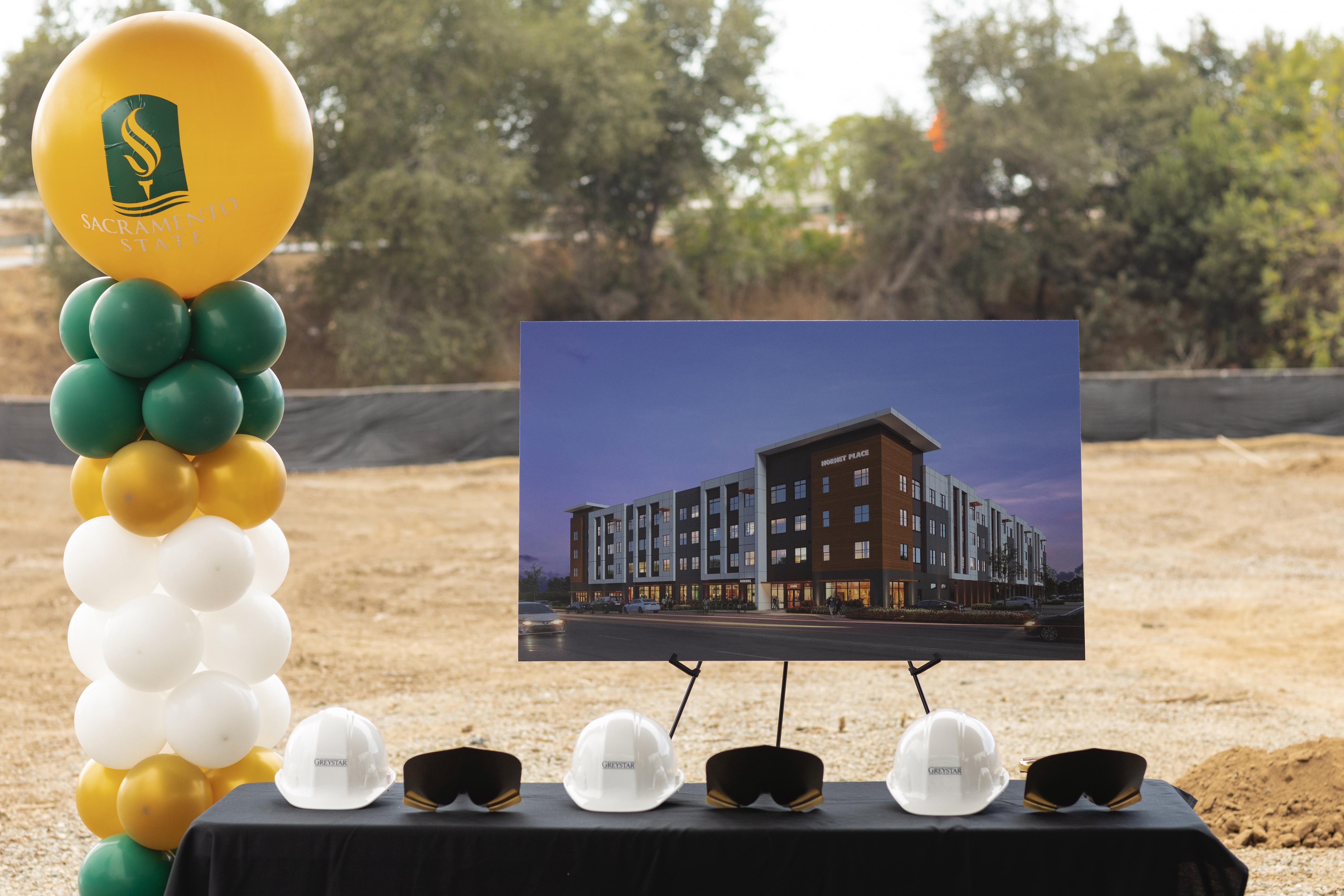 Hardhats and shovels sit on a table flanked by green and gold balloons with a large drawing of a new housing complex.