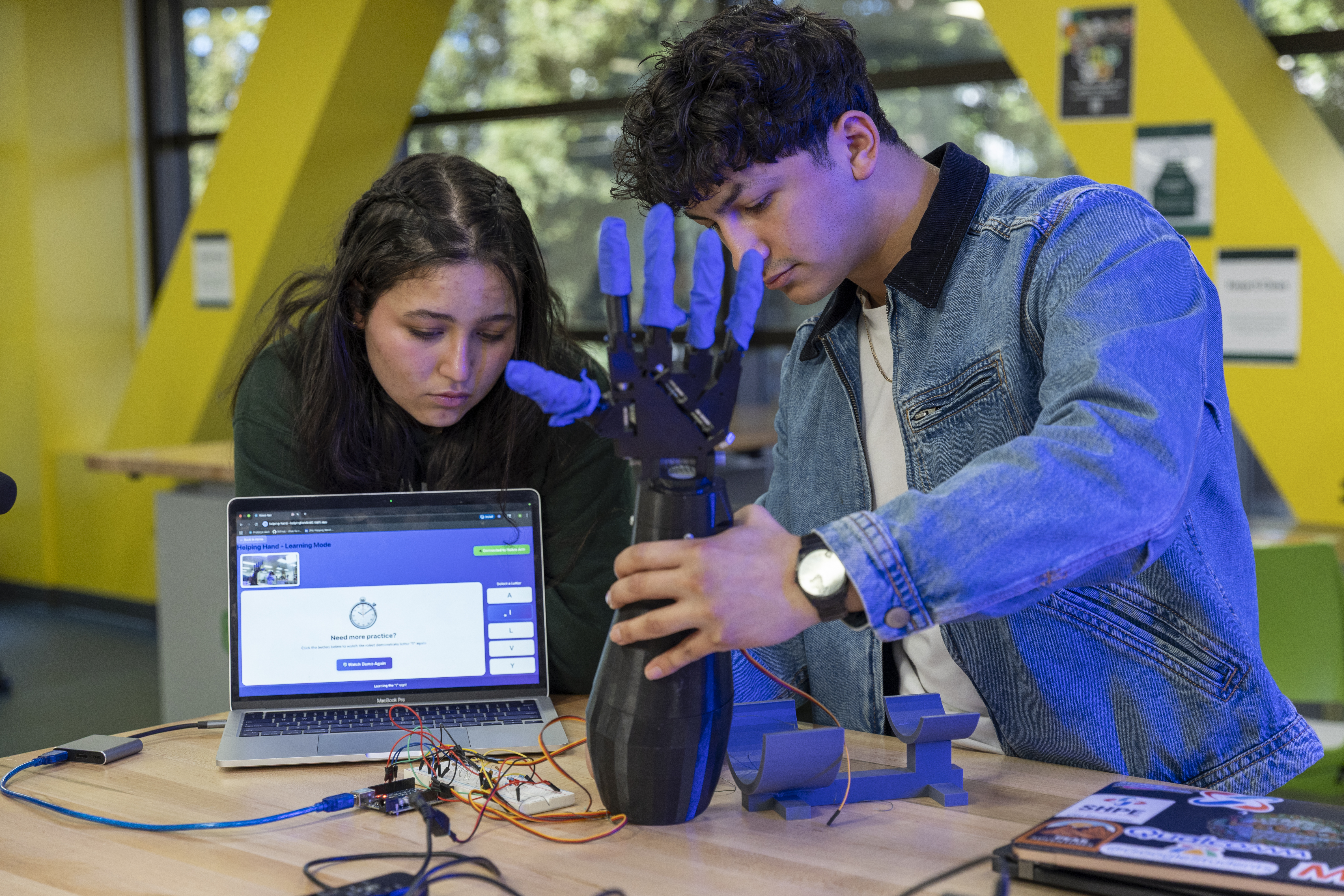A woman and man work over a laptop computer while holding a robotic arm.