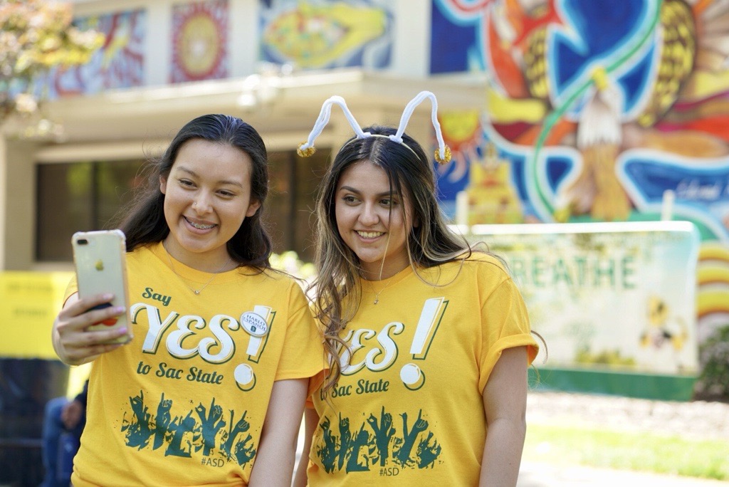 Two women in yellow t shirts pose for selfies during Admitted Student Day