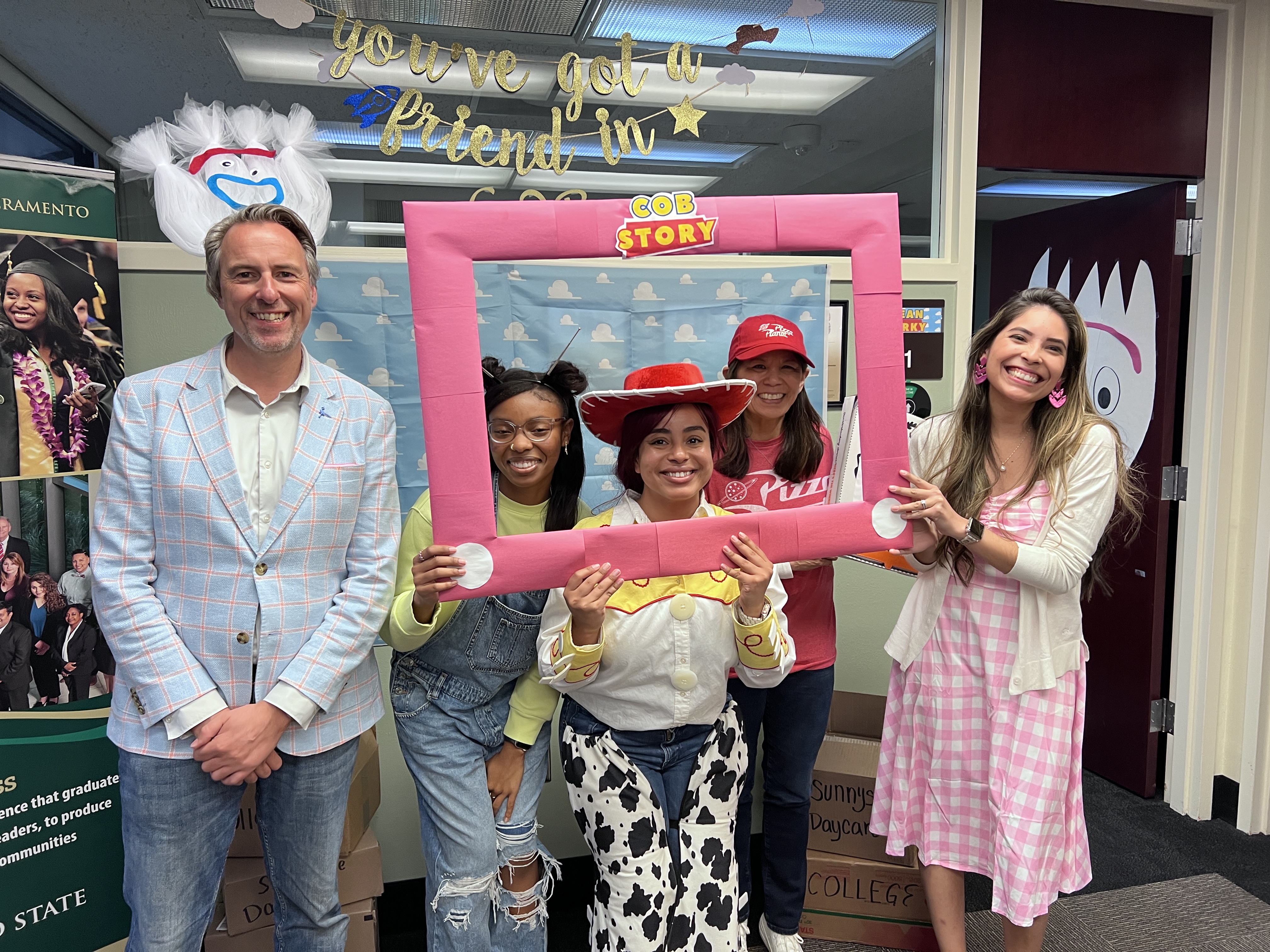 College of Business Dean’s Office: C.O.B. Story group dressed in costumes posing behind a COB picture frame in front of decorated Toy Story theme office