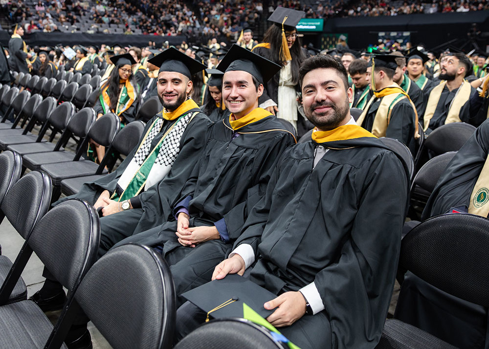 Three male students sitting and smiling at the camera during graduation ceremony with their cap and gowns.