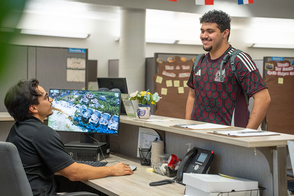 A student assistant greeting with a smile another student behind a desk.
